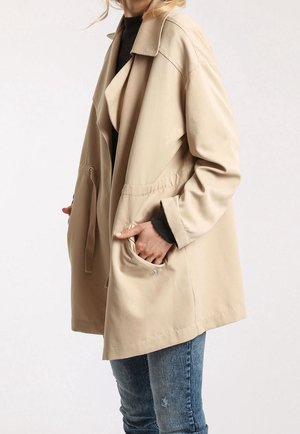 Persoon in een beige trenchcoat over een donkere top, met hand in de zak, gecombineerd met blauwe versleten spijkerbroek tegen een effen achtergrond.