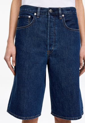 Osoba nosząca ciemnoniebieskie denimowe spodenki za kolano z przednimi kieszeniami i zapinane na guziki, stojąca na jednolitym białym tle.