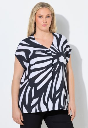 Blonde Frau, die eine lockere, schwarz-weiße Bluse mit abstraktem Muster und kurzen Ärmeln trägt, vor einem schlichten hellen Hintergrund steht.