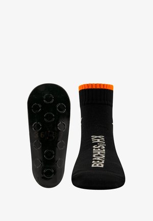 Chaussette noire avec bord côtelé orange, fond texturé avec des grips circulaires, et logo imprimé en blanc sur le côté.