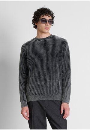 Giovane uomo che indossa un maglione a coste scuro, pantaloni neri e occhiali da sole rettangolari, guarda leggermente di lato su uno sfondo semplice.