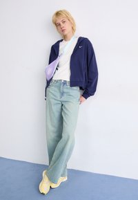 Marineblauwe rits hoodie, witte t-shirt, lichtblauwe wijde jeans, gele sneakers en een kleine paarse schoudertas tegen een neutrale achtergrond.