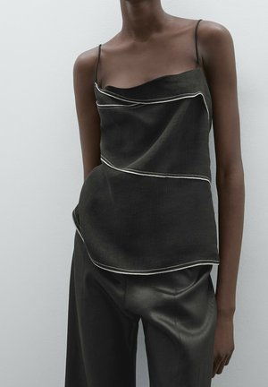 Ciemny bezrękawnik z białymi lamówkami noszony z luźnymi czarnymi spodniami na osobie stojącej na prostym jasnym tle.