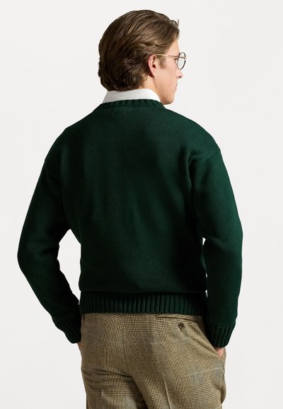 Donkergroen gebreide trui met een geribde zoom en manchetten, met een ronde hals, gedragen over patroonbeige broek.