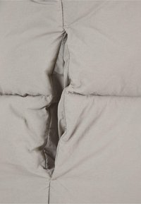 Siva puffer jakna sadrži preklopni dizajn s vidljivim šavovima, mekanom teksturom i malim otvaranjem duž šava za dodatnu izolaciju.