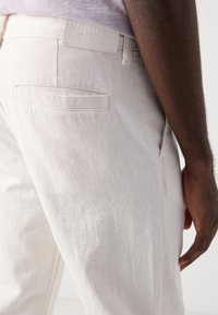 Beiga linbyxor med vävd textur, som har en bakficka och en vit märkesetikett vid midjan.