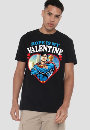 Young man wearing black T-shirt with Superman inside a heart and text "Hope is my Valentine" against cityscape background.