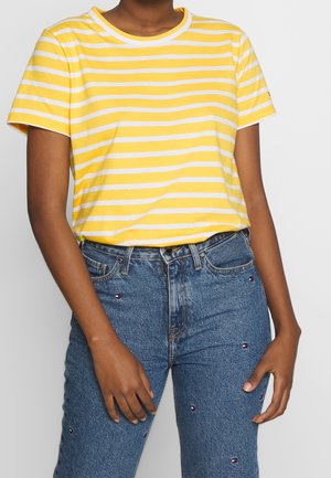 Personne portant une chemise à manches courtes rayée jaune et blanche et un jean en denim bleu avec de petits logos brodés.