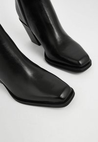Stivaletti neri in pelle con punta quadrata e tacco a blocco, caratterizzati da una texture liscia e una leggera lucentezza. Chiusura con zip sul lato.