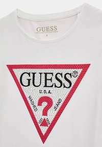 Weißes Baumwoll-T-Shirt mit einem großen roten dreieckigen Logo, auf dem "GUESS" und "USA" in Schwarz steht, umgeben von kleinen Nieten und einem Fragezeichen.