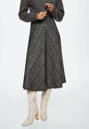 Ubrana w szerokie culottes w czarno-białą kratę, brązowy krótki sweter i kremowe kozaki za kolano na obcasie, ręce blisko brzucha.