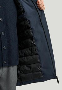 Marinblå jacka med dragkedja, med en slät ytteryta och vadderad svart foder. Innehåller en knappad cardigan under.