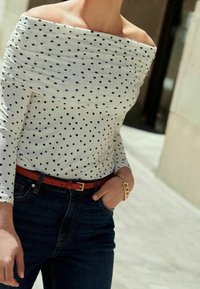 Top bianco con spalle scoperte e pois neri, abbinato a jeans in denim scuro e una cintura rossa. Bracciale in catena dorata visibile al polso.
