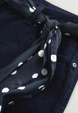 Marineblauer Cord-Stoff mit strukturierter Oberfläche, versehen mit einem gebundenen marineblauen und weißen Seidengürtel mit Punkten an der Taille, akzentuiert durch Knöpfe.