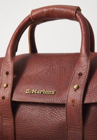Barna bőr táska, texturált felülettel, két felső fogantyúval, aranyszínű "Dr. Martens" logóval és sárgaréz kiegészítőkkel.