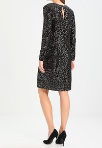 Robe noire à paillettes avec des manches longues, un détail de dos en tour de clé et une silhouette ajustée, arrivant au-dessus du genou. Portée avec des talons noirs.
