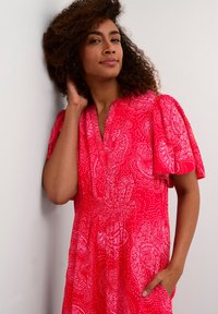 Červené šaty s paisley vzorem a bílými akcenty. Mají krátké balónkové rukávy, výstřih ve tvaru V a dvě boční kapsy. Vyrobeny z lehkého materiálu.