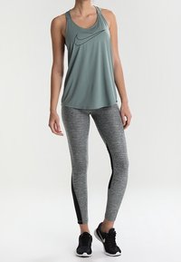 Top senza maniche di colore verde chiaro con logo nero abbinato a legging grigi con un accento nero sul lato; scarpe da ginnastica nere completano l'outfit.