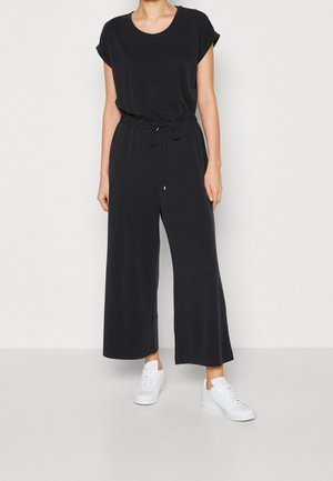 Musta haalaripuku, jossa on lyhyet hihat, pyöreä kaula-aukko ja kiristysnauha vyötäröllä. Laskoshousut ja kevyt kangas, yhdistettynä valkoisiin lenkkareihin.