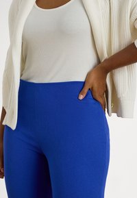 Nő fehér ujjatlan felsőt, krémszínű kötött kardigánt és élénk kék, testhezálló nadrágot visel, egyik kezét a csípőjén pihentetve.