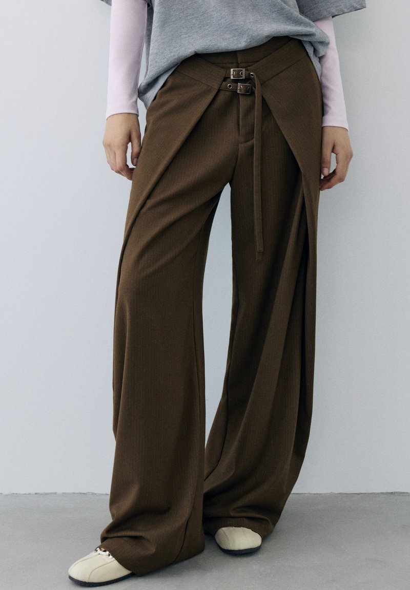 Pantalon marron à jambes larges avec panneaux avant superposés maintenus par deux petites boucles, porté avec des chaussures blanches et des hauts superposés.