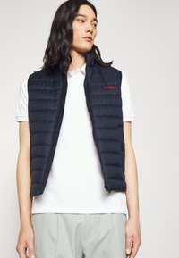 Chaleco acolchado navy con diseño de acolchado vertical, cuello alto y logo rojo "HUGO" en el pecho. Con cremallera frontal y diseño sin mangas.