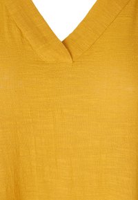 Senapsgul topp med V-ringning, tillverkad av texturerat tyg, med subtila sömdetaljer överallt.