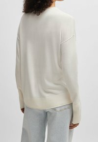 Frau mit lockigem Haar, die einen lockeren weißen Pullover und hellblaue Jeans trägt, von hinten vor einem schlichten Hintergrund betrachtet.