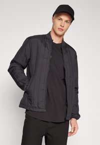 Giacca bomber nera trapuntata con chiusura a zip, colletto a coste e polsini elasticizzati. Indossata sopra una maglietta nera e pantaloni neri. Berretto sullo sfondo.