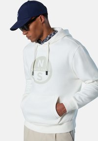 Felpa bianca con cappuccio con un grande logo circolare grigio, tasca frontale, coulisse e polsini a costine; indossata sopra una camicia a quadri, abbinata a un berretto blu navy.