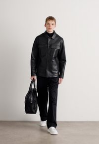 Chaqueta de camisa de cuero negro con dos bolsillos en el pecho, combinada con pantalones negros y zapatillas blancas, sostenida con un bolso negro en la mano.
