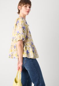 Blumen-Peplum-Bluse in Gelb, Lila und Grün mit puffartigen Ärmeln, kombiniert mit dunkelblauen Jeans und einer gelben Handtasche. Weicher, leichter Stoff.