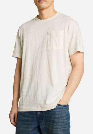 Homme portant un T-shirt texturé à manches courtes de couleur écrue avec une poche poitrine, associé à un jean bleu, main dans la poche, fond neutre.