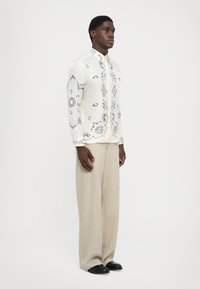 Weißes Hemd mit blauen Blumenmustern, Stehkragen, Knopfverschluss, kombiniert mit hellbeigen, weit geschnittenen Hosen und schwarzen Schuhen.