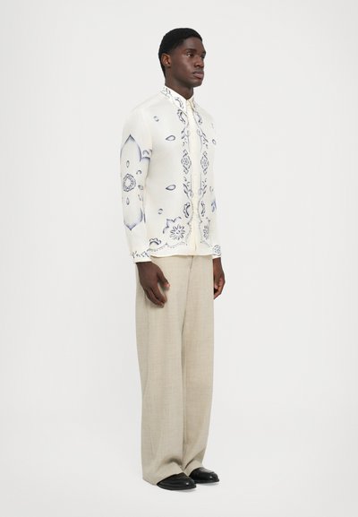 Valkoinen paita sinisillä kukkakuoseilla, pystykaulus, nappikiinnitys, yhdistetty vaaleanbeigeihin leveälahkeisiin housuihin ja mustiin kenkiin.