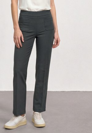 Femme portant un pantalon droit gris foncé avec poches plaquées à l’avant et des baskets blanches, debout devant un fond clair uni.