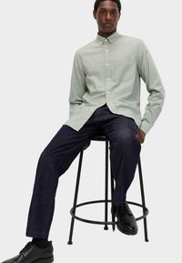 Camicia verde chiaro con bottoni e taschino sul petto, abbinata a jeans blu scuro e scarpe nere, seduta su uno sgabello in metallo nero.