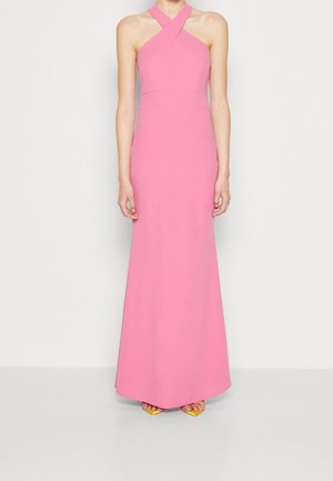 Kobieta ubrana w różową, sięgającą podłogi suknię na halter z krzyżującymi się ramiączkami, stojąca na jednolitym białym tle.