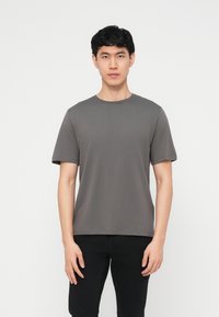 Young man standing against white background wearing a plain grey short-sleeve t-shirt and black pants, facing forward with neutral expression.