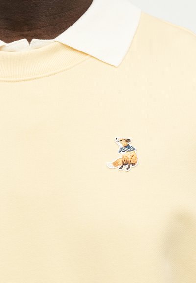 Ανοιχτόκίτρινο πουλόβερ με λευκό γιακά και μικρή κεντημένη αλεπού που φοράει μπλε κασκόλ στο στήθος.