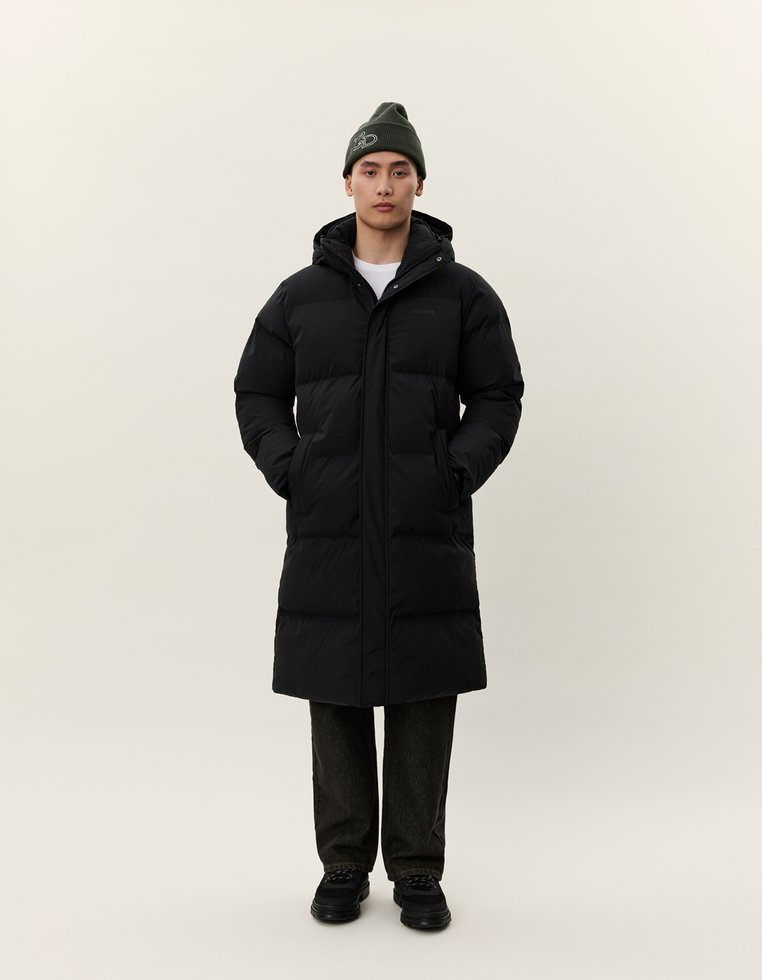 Uomo in piedi, che indossa un lungo cappotto imbottito nero con cappuccio, pantaloni scuri, scarpe nere e un berretto verde con un design bianco.