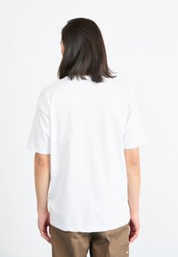 Weißes Baumwoll-T-Shirt mit kurzen Ärmeln, gekennzeichnet durch einen Rundhalsausschnitt und einen lässigen Schnitt, von hinten gesehen. Kombiniert mit braunen Hosen.