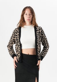 Eläinkuosilla varustettu beigen ja mustan värinen neuletakki, jossa on pitkät hihat, napit ja ribbireunat hihoissa ja helmassa. Päällä valkoinen cropped-top.