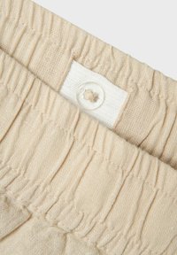 Tissu beige à texture plissée, avec une étiquette blanche et un détail de bouton transparent. Matériau lisse, finition douce et bords cousus.