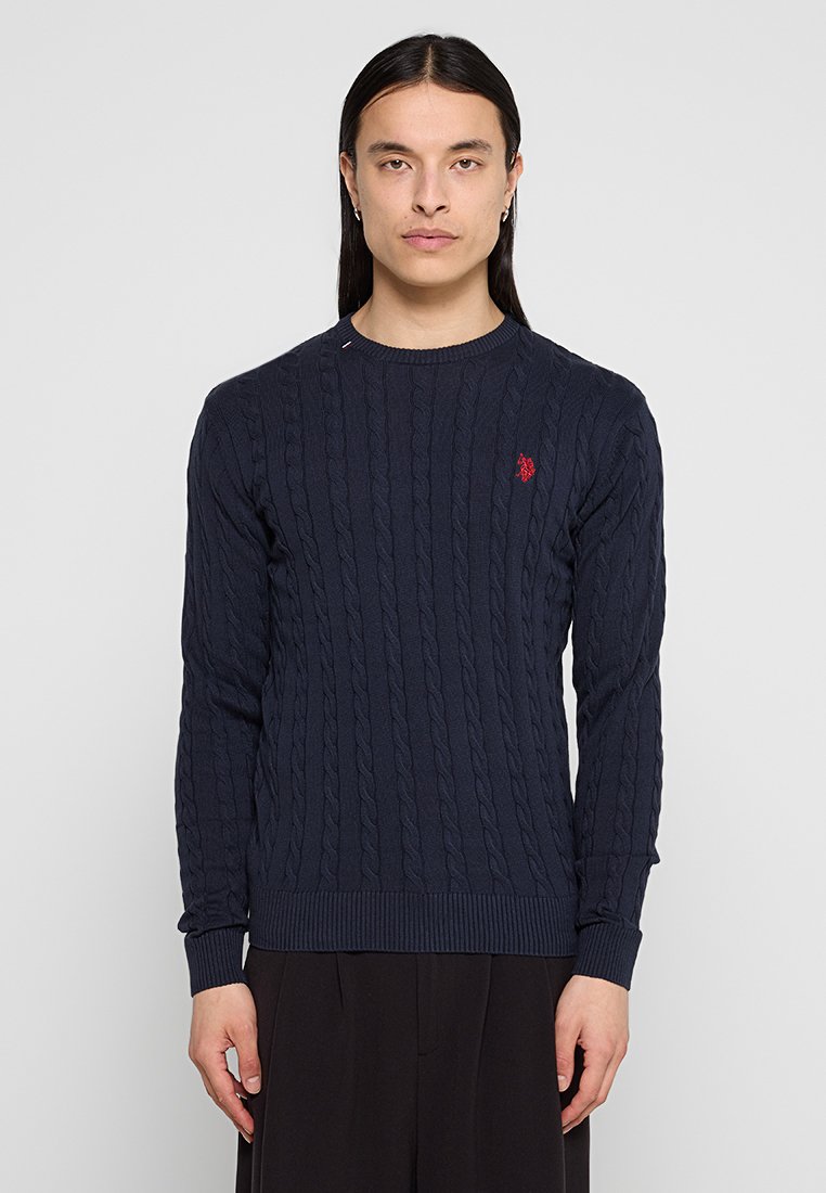 Junge Person mit langen dunklen Haaren, die einen navyfarbenen Kabelstrickpullover mit rotem besticktem Logo und schwarze Hosen trägt, steht vor einem schlichten Hintergrund.