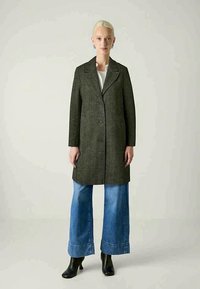 Naine, kes kannab tumedat halli herringbone-stiilis mantelil valget topsi, laia jalgadega siniseid teksaseid ja musti pahkluu saapaid, seisab ühetoonilise taustaga.