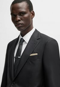 Czarny garnitur o teksturowym wykończeniu, biała koszula i ciemny krawat z delikatnymi paskami. Wyposażony w srebrny klips do krawata i wzorzystą chusteczkę w kieszeni.