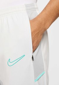 Nike shorts i hvid med en grå meshpanel, der har en sidelomme med lynlås og et broderet turkist logo. Glat stoftekstur.