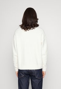 Bílý svetr s kulatým výstřihem a žebrovaným lemem, vyrobený z měkkého materiálu, ve spojení s tmavě modrými džíny.