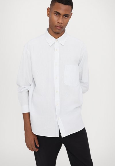 Camisa blanca de manga larga con sutiles rayas verticales, con botones en el frente y un único bolsillo en el pecho. Combinada con pantalones oscuros.